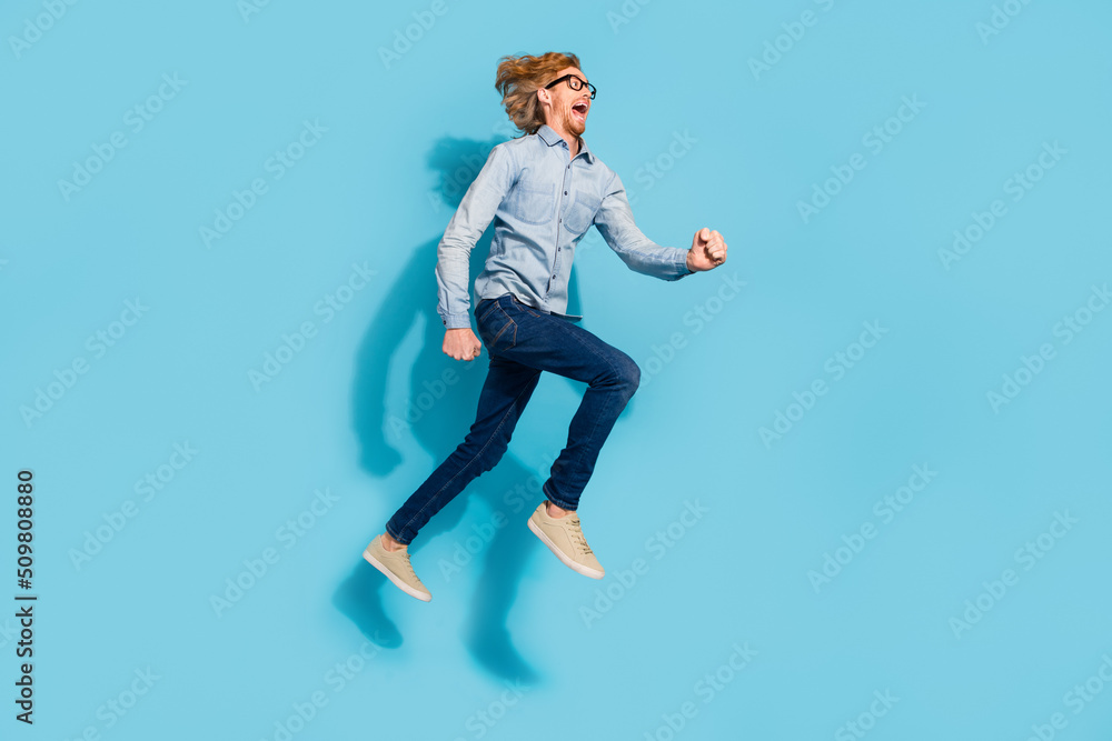 Full length photo of shocked excited young guy dressed jeans shirt eyewear jumping high running fast isolated blue color background