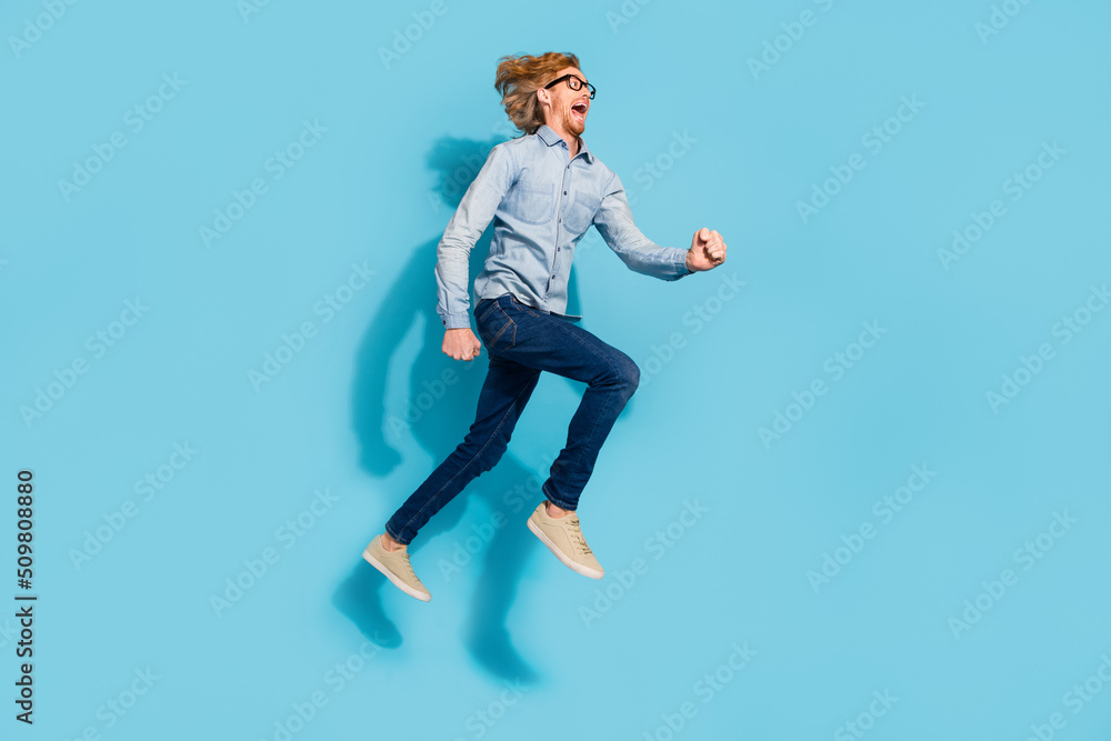 Full length photo of shocked excited young guy dressed jeans shirt eyewear jumping high running fast isolated blue color background Full length photo of shocked excited young guy dressed jeans shirt eyewear jumping high running fast isolated blue color background