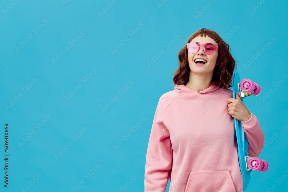 Funny joyful pretty redhead lady in pink hoodie sunglasses with penny board smiling posing isolated on blue studio background. Copy space Banner Offer. Fashion Cinema. Holiday activity Funny joyful pretty redhead lady in pink hoodie sunglasses with penny board smiling posing isolated on blue studio background. Copy space Banner Offer. Fashion Cinema. Holiday activity