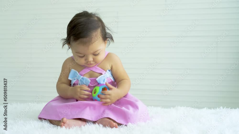 Adorable caucasian baby girl in pink clothes sitting on bed at home, looking away, playing toy color ball. Kid at home and childhood concept Adorable caucasian baby girl in pink clothes sitting on bed at home, looking away, playing toy color ball. Kid at home and childhood concept