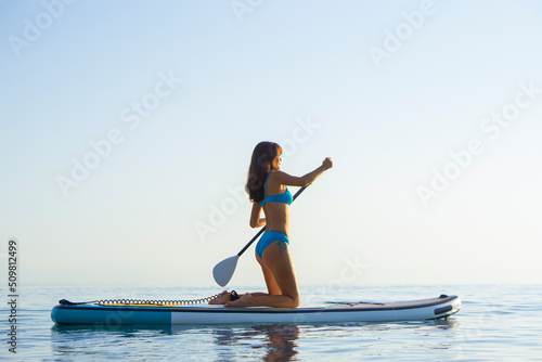 Asian beautiful girl in a blue bikini is engaged in water sports standing surfing paddle Sup in the ocean on the background of sunset. Woman standing up paddleboarding.
