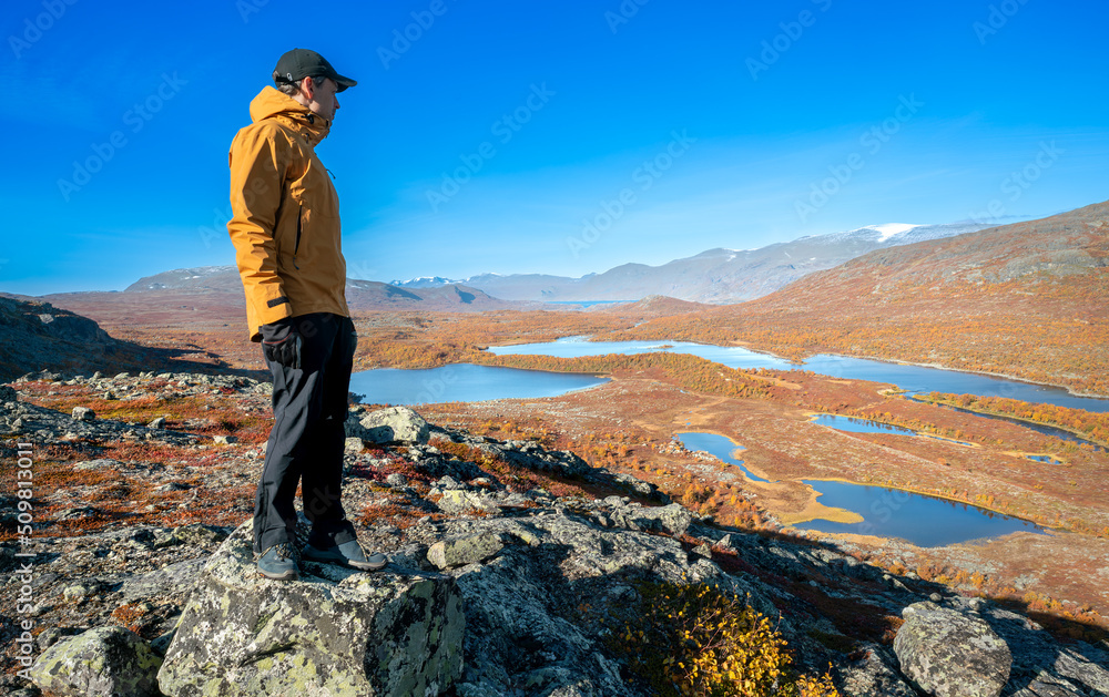 Obraz premium Male hiker in yellow hardshell jacket and black pants overlooking epic view of vast arctic landscape of Stora Sjofallet National Park, Sweden, on autumn day. Mountains and valleys of Lapland.