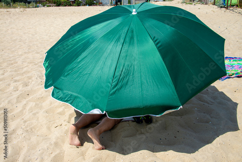 A green umbrella on a sandy beach. Legs of a vacationer look out from under an umbrella on a sandy sea beach.