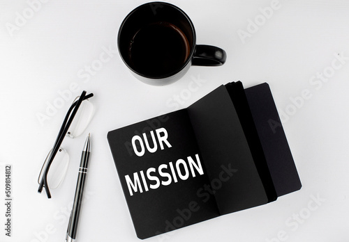 OUR MISSION written text in small black notebook with coffee , pen and glasess on white background. Black-white style