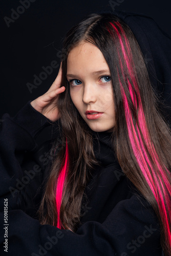 portrait teenage girl with dark hair with pink strands  dressed in fashionable clothes  hoodie with hood  stands on black background and looks at camera. transitional age. concept youth and clothing