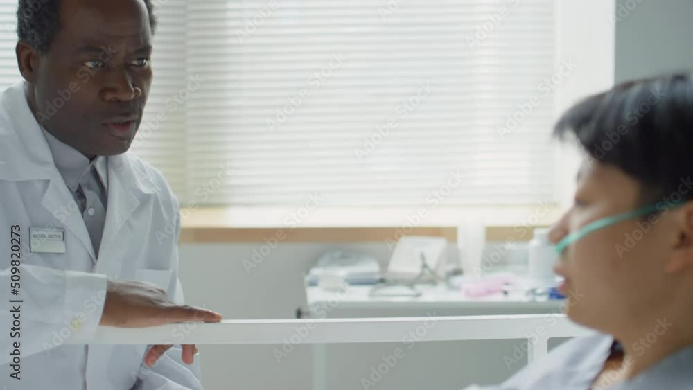 Rack focus shot of African American doctor sitting by bed in hospital ward and having discussion with Asian female patient while visiting her during therapy Rack focus shot of African American doctor sitting by bed in hospital ward and having discussion with Asian female patient while visiting her during therapy