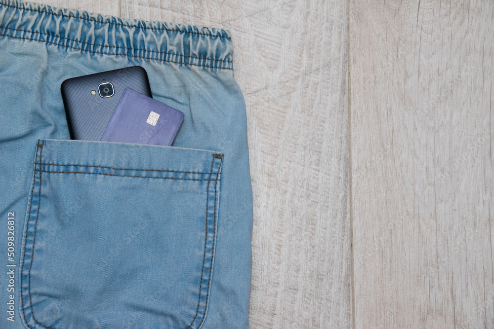 Bank card and smartphone in light blue jeans pocket on light wooden background with copy space Bank card and smartphone in light blue jeans pocket on light wooden background with copy space