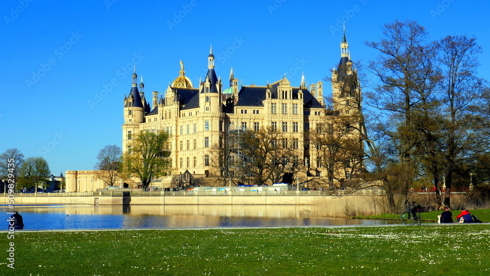 prächtiges Schloss in Schwerin am Burgsee neben Schilf und Bäumen hinter grüner Wiese unter blauem Himmel