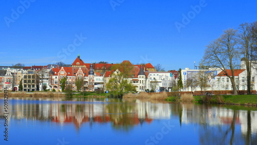 schöner Blick über spiegelnden Burgsee auf Häuser in Schwerin mit Bäumen unter blauem Himmel