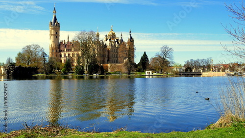 prächtiges Schloss in Schwerin am See neben Schilf und Bäumen hinter grüner Wiese unter blauem Himmel