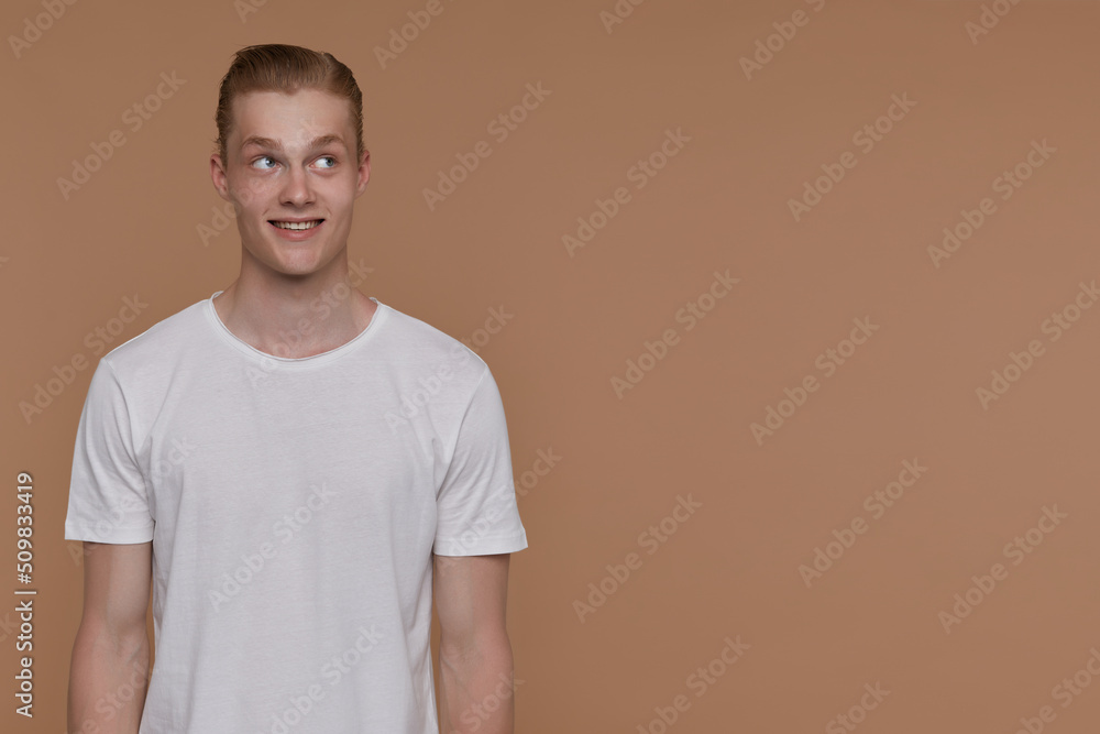 Indoor portrait of young blonde man posing over beige background looking aside at copy space with smiling and confused facial expression Indoor portrait of young blonde man posing over beige background looking aside at copy space with smiling and confused facial expression