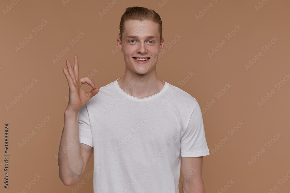 Indoor portrait of young blonde man posing over beige background showing correct sign into camera and smiling broadly Indoor portrait of young blonde man posing over beige background showing correct sign into camera and smiling broadly