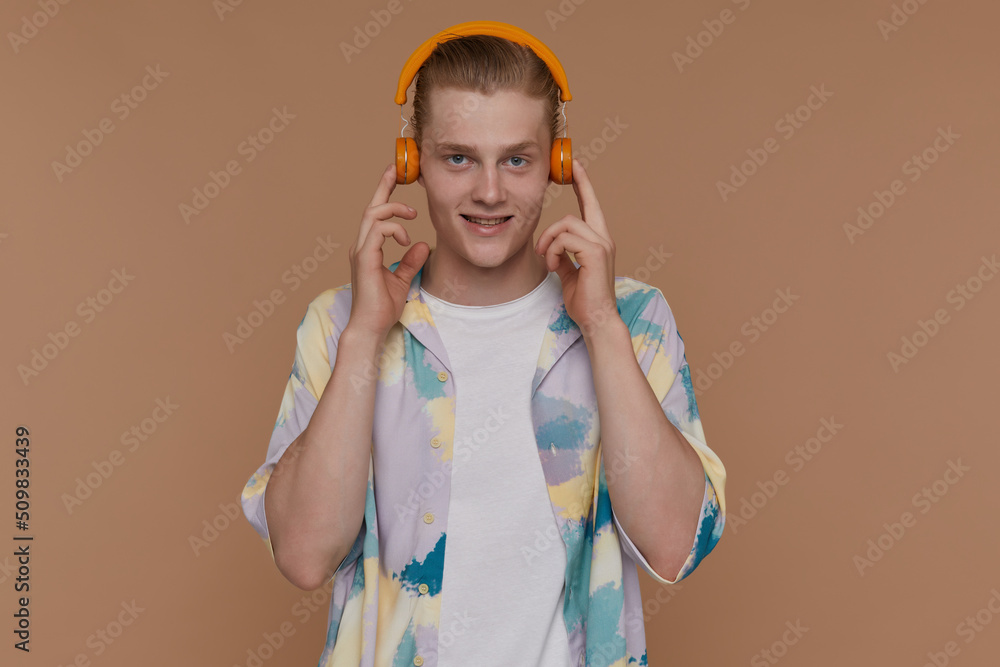 Indoor portrait of young blonde man posing over beige background looking into camera while listening music Indoor portrait of young blonde man posing over beige background looking into camera while listening music