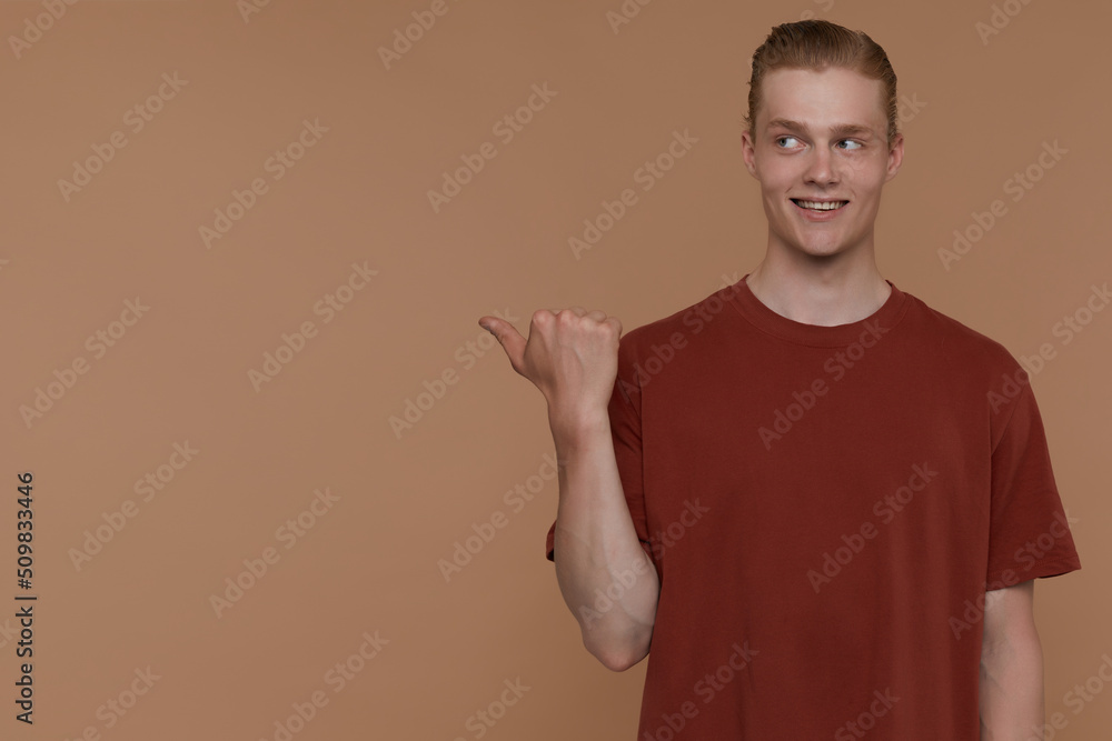 Indoor portrait of young blonde man posing over beige background points with a finger aside at copy space and smiles broadly Indoor portrait of young blonde man posing over beige background points with a finger aside at copy space and smiles broadly