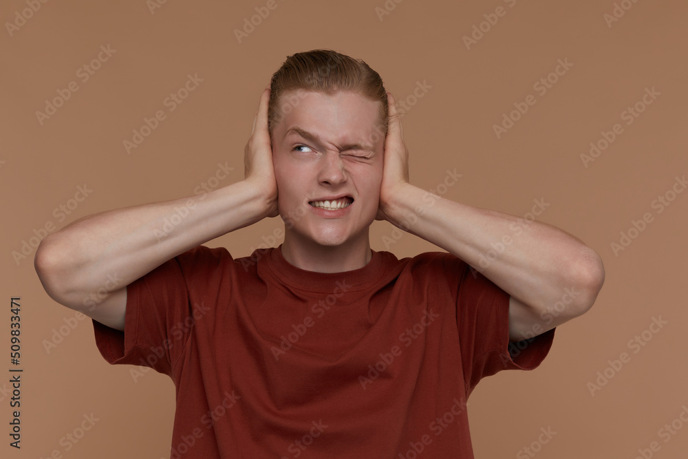 Indoor portrait of young blonde man posing over beige background close his ears with palms Indoor portrait of young blonde man posing over beige background close his ears with palms