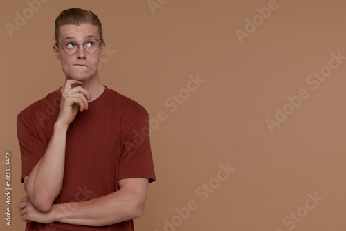 Indoor portrait of young blonde man posing over beige background biting his lip and looks aside at copy space © timtimphoto