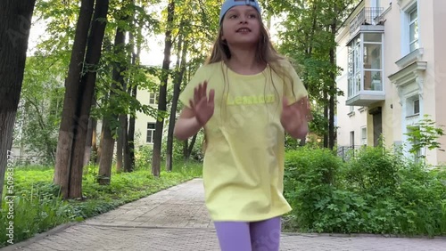 A beautiful little girl in a yellow t-shirt and a blue cap dances on the street in the park, freedom of movement, happy childhood photo