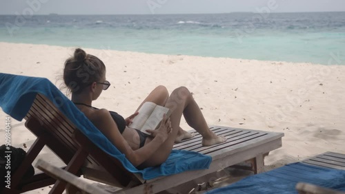 An attractive girl in a swimming suit sits on sun lounger on the beach with a book in her hands. A calm blue sea ahead.