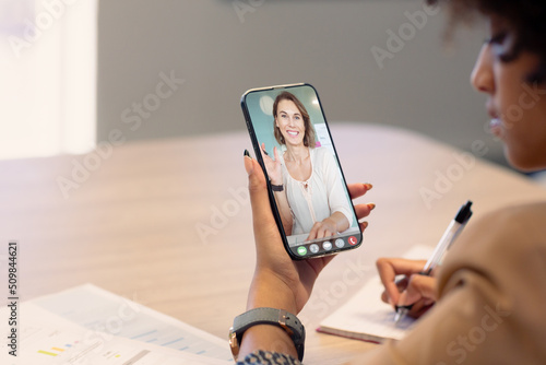 Biracial businesswoman making notes while video calling caucasian female colleague on smart phone