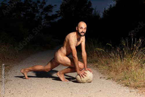 Young male athlete fully naked crouching on a trail at night looking at camera