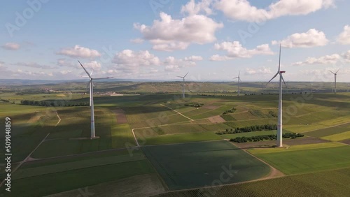 Numerous tall wind turbines standing within lush green meadows and grapevine fields underneath a blue sky with cumulus clouds. Wide angle aerial approach photo