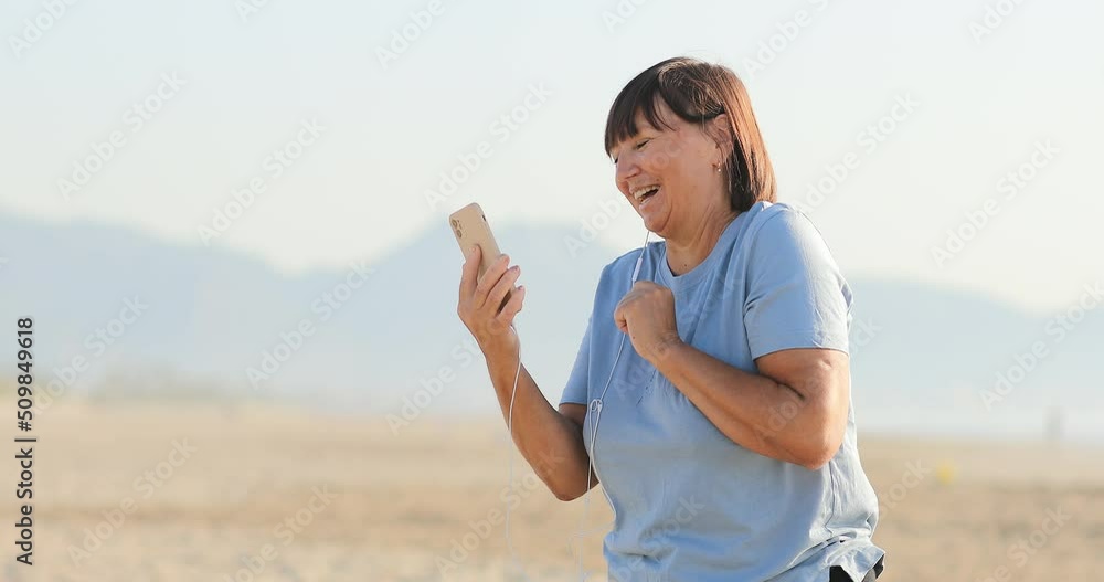 smiling middle aged woman talking on video call using smartphone with headphones on the beach. female 50 years remote has conversation with family on vacation. smiling middle aged woman talking on video call using smartphone with headphones on the beach. female 50 years remote has conversation with family on vacation.