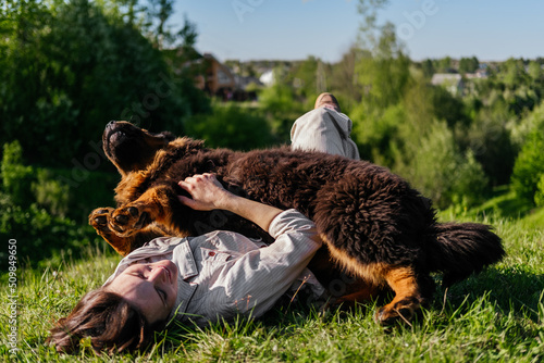 Laughing woman in casual clothes laying on grass and playing with tibetan mastiff dog outdoors