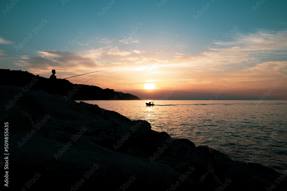 Fototapeta premium SILHOUETTE OF FISHERMEN AT SUNSET ON THE CLIFFS BY THE SEA WITH BOAT IN THE BACKGROUND