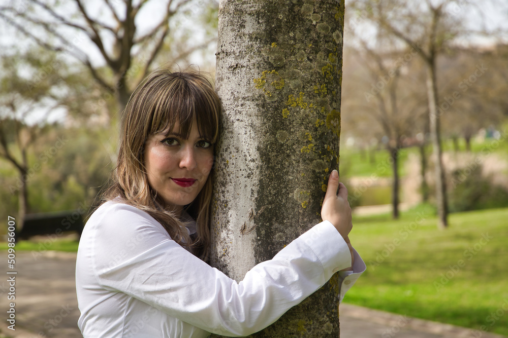 Young, beautiful, blonde woman in white shirt, hugging a tree in the middle of a park. Concept beauty, nature, love, climate change, naturalist.