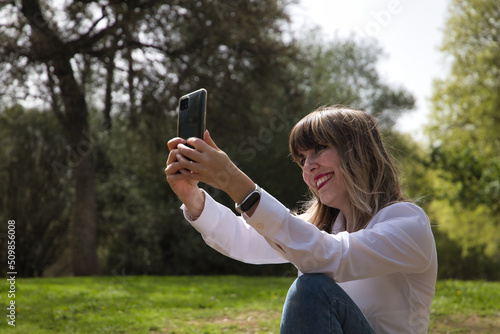 Portrait of young, beautiful, blonde woman in white shirt and jeans, taking a selfie with her cell phone in the middle of a park. Mobile concept, selfie, photo, portrait, technology.