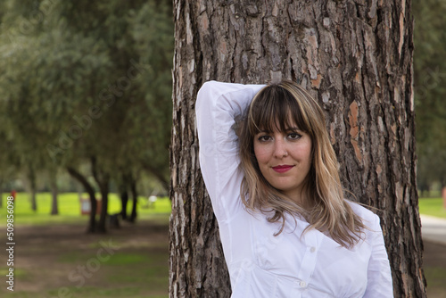 Fotografia Portrait of young, beautiful, blonde woman in white shirt leaning against the trunk of a large tree