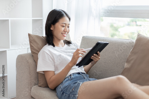 Asian woman sitting on the sofa in the living room using a tablet, Looking at tablet screen, Relaxing at home, Working through the Internet communication system, Happy working lifestyle.