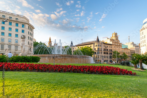 Photography Fountain with red flowers in the foreground at the Plaza de Catalunya in Barcelona , Spain as the sun begins to set in the distance and the city is in view