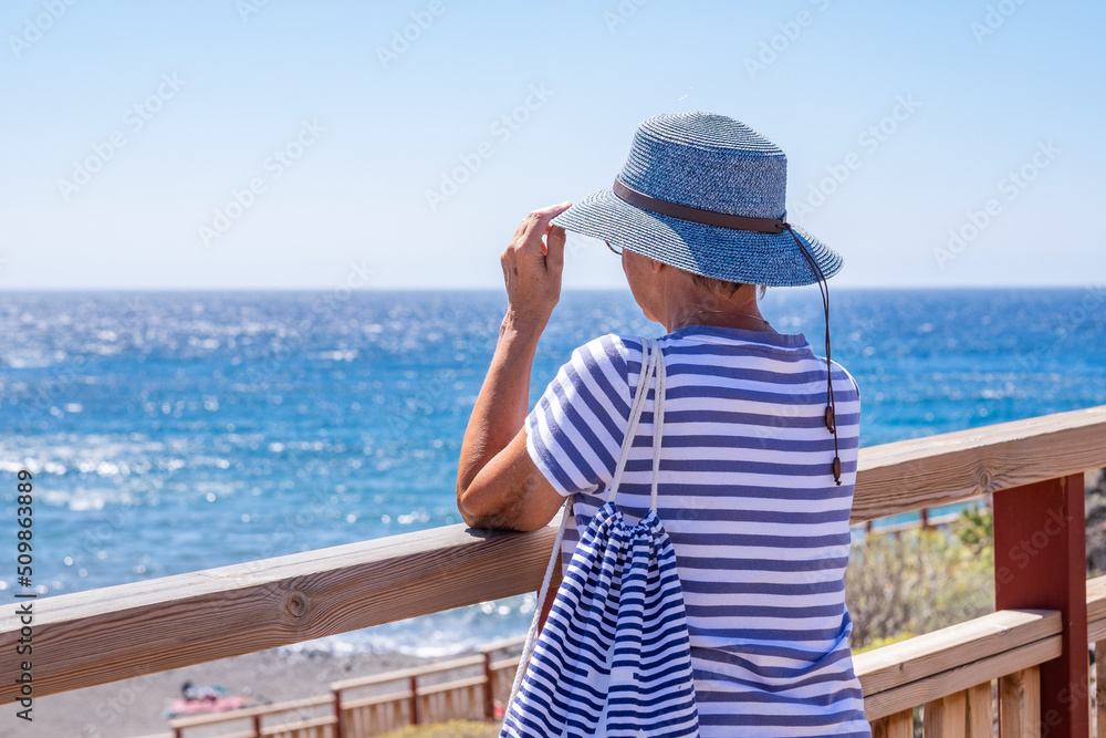Rear view of mature woman dressed in blue admiring the sea in a summer holiday looking at horizon over water. Mature lady enjoying relaxation and free time