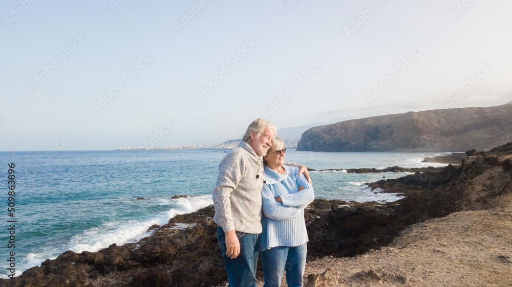 Senior caucasian couple at the sea hugging each other on the cliff watching the sunrise enjoying freedom, vacation or retirement