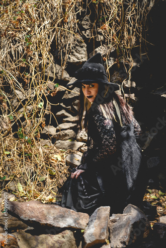 Beautiful young, gothic, sexy and witch woman from with stylized hands with black nails and accessories, magic wooden wand and witch hat in the forest between the nature © Samuel Ponce