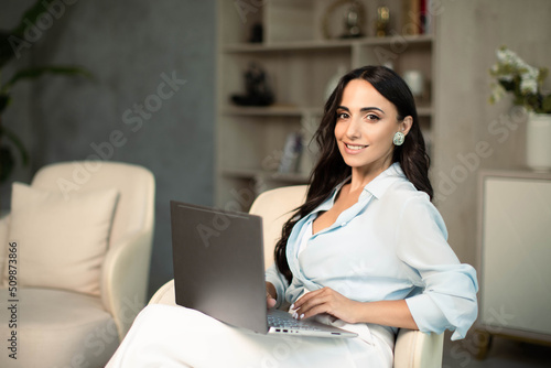 Woman sitting on chair indoors at home apartment, using laptop computer, self employed remote work and small business freelancer, education and leisure time, online shopping, communication technology