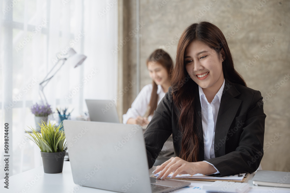 Young business women working on laptop with happy and smile face with co-worker is talking via mobile phone behind her in office spec. Young business women working on laptop with happy and smile face with co-worker is talking via mobile phone behind her in office spec.