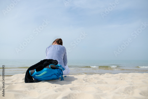 Dziewczyna, kobieta z plecakiem siedzi na piaszczystej morskiej plaży. Wpatruje się w horyzont. Lato, pastelowe kolory, przydymione błękitne niebo.