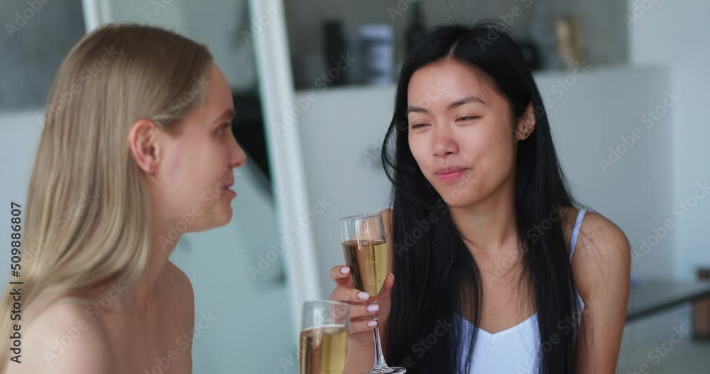 Close-up of two cheerful attractive blonde and brunette women chilling together at hen-party , drinking sparkling wine from a glass sitting at bathtub at home bathroom. Focus on Asian pretty woman Close-up of two cheerful attractive blonde and brunette women chilling together at hen-party , drinking sparkling wine from a glass sitting at bathtub at home bathroom. Focus on Asian pretty woman