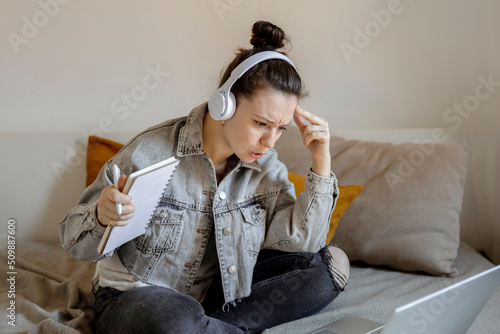 Angry and dissatisfied woman with casual clothes sitting on bed at home with computer and studying. Girl is sad and tired. Negative emotions, stress, mental problems, deadline. Distance education. photo