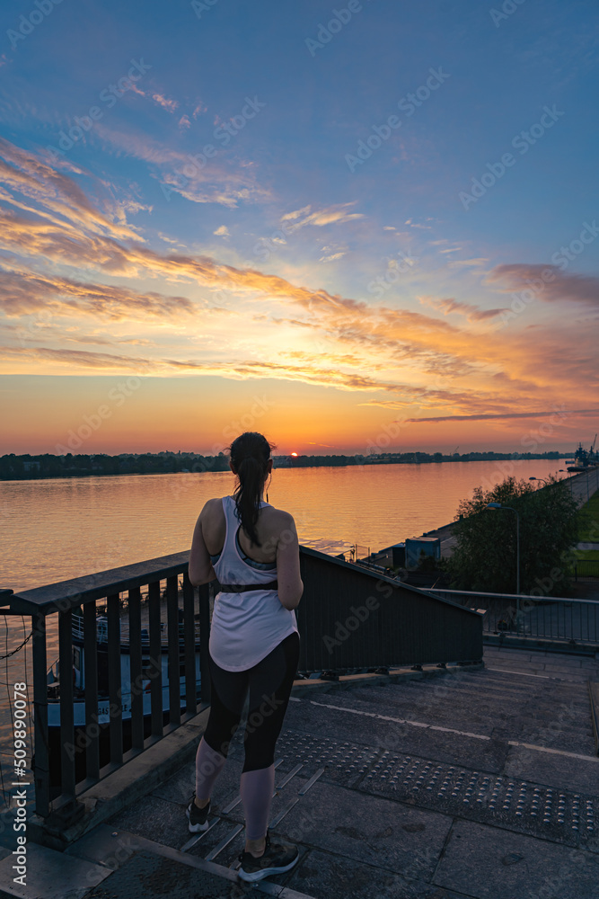 Fototapeta premium Woman in the sportswear on the bridge watching stunning sunset over the river