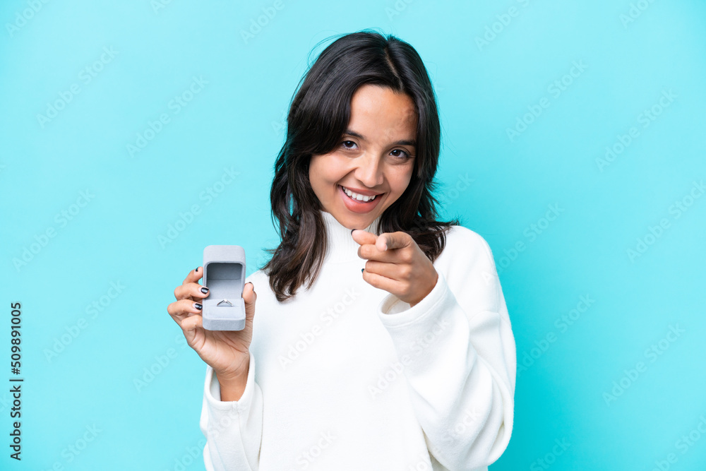 Young hispanic woman holding a engagement ring isolated on blue background pointing front with happy expression Young hispanic woman holding a engagement ring isolated on blue background pointing front with happy expression