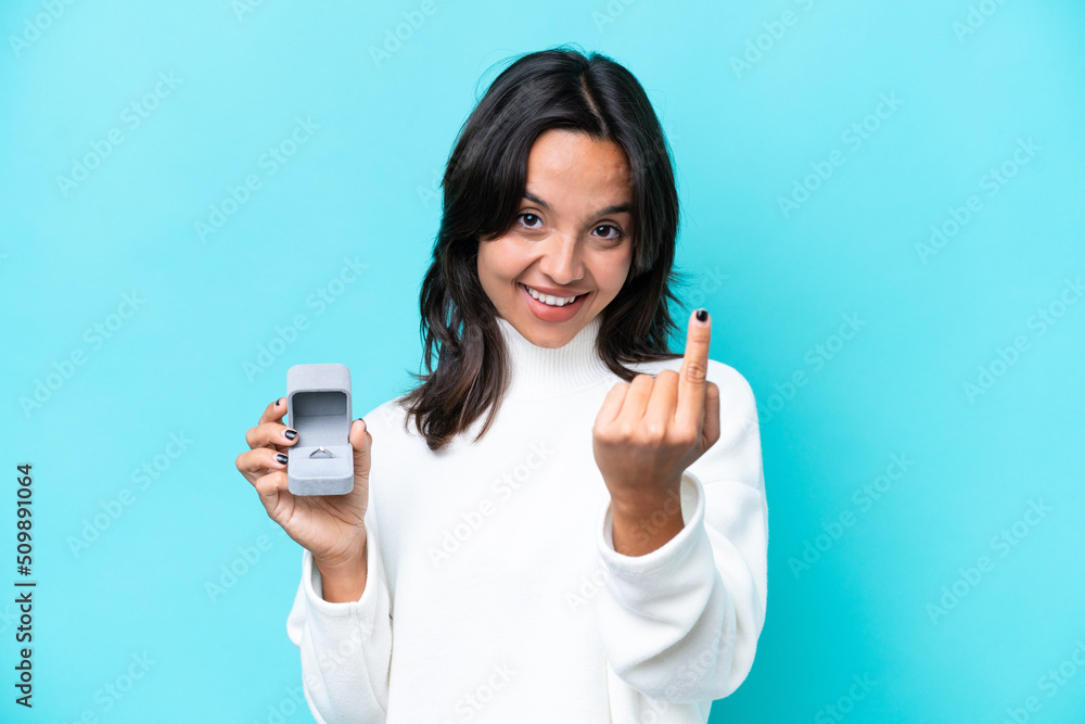 Young hispanic woman holding a engagement ring isolated on blue background doing coming gesture Young hispanic woman holding a engagement ring isolated on blue background doing coming gesture