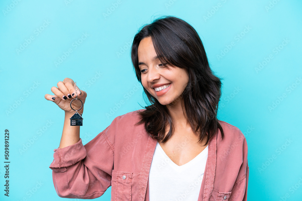 Young hispanic woman holding home keys isolated on blue background looking to the side and smiling Young hispanic woman holding home keys isolated on blue background looking to the side and smiling