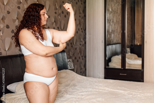 Curly plus size young woman in white underwear stand near bed in bedroom and pinch sagging arms by hand side view. Overweight girls struggle with obesity. Weight loss program, body control and diet.