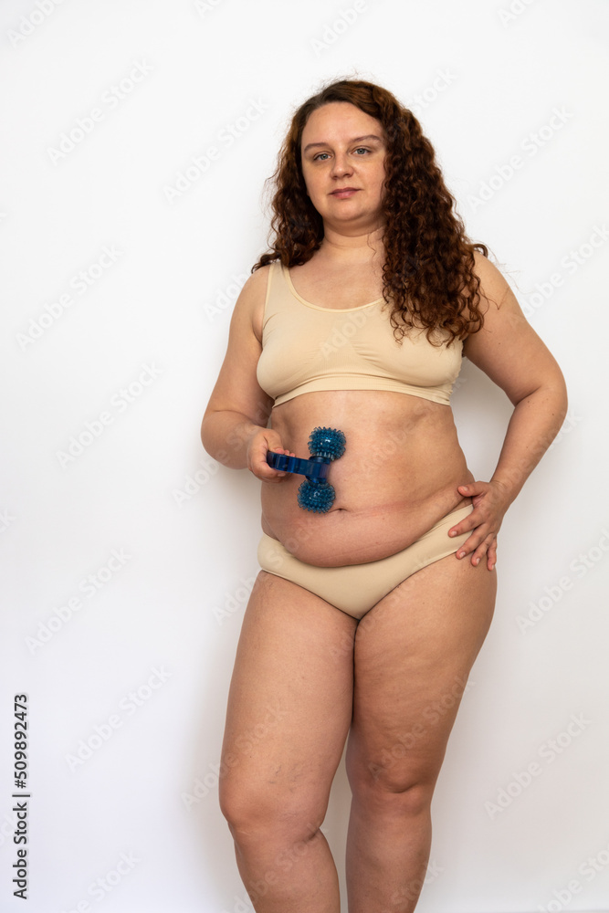 Obraz premium Portrait of serious fat middle-aged woman with long curly hair wearing beige underwear, massaging flabby belly with blue plastic roller, standing on white background. Weight loss, lymphatic drainage.