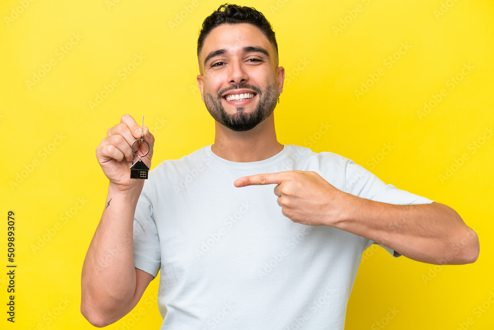 Young Arab man holding home keys isolated on yellow background and pointing it Young Arab man holding home keys isolated on yellow background and pointing it