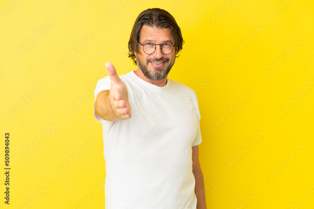 Senior dutch man isolated on yellow background shaking hands for closing a good deal Senior dutch man isolated on yellow background shaking hands for closing a good deal