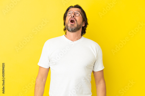 Senior dutch man isolated on yellow background looking up and with surprised expression